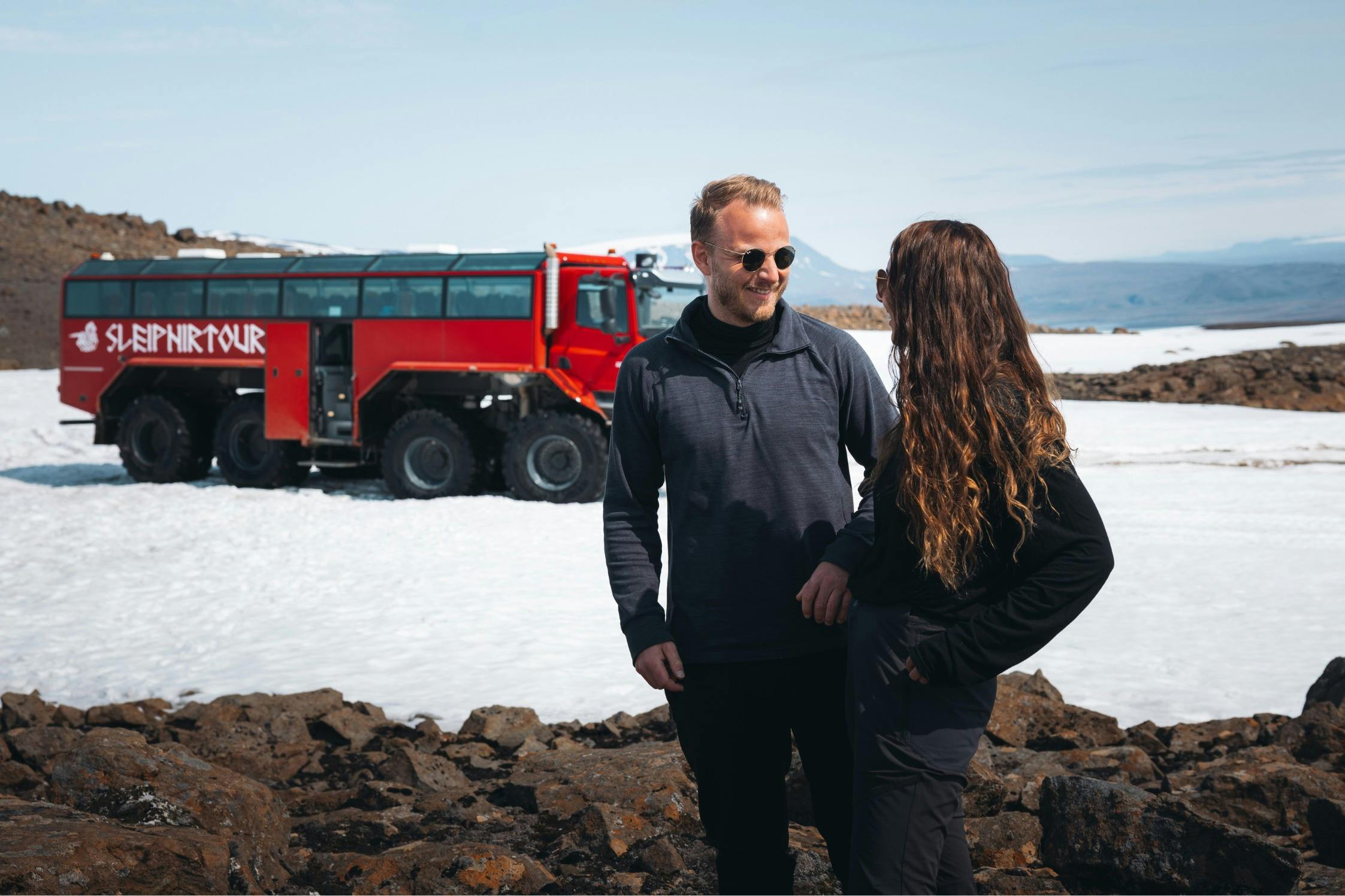Zwei Menschen stehen auf felsigem Gelände mit einem roten Expeditionsfahrzeug im Hintergrund, Schnee und Berge sind zu sehen.