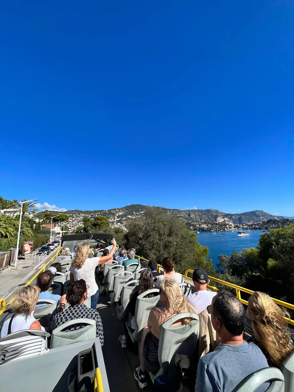 People seated on an open-top tour bus overlooking a coastal town with hills, trees, and a vivid blue sky.