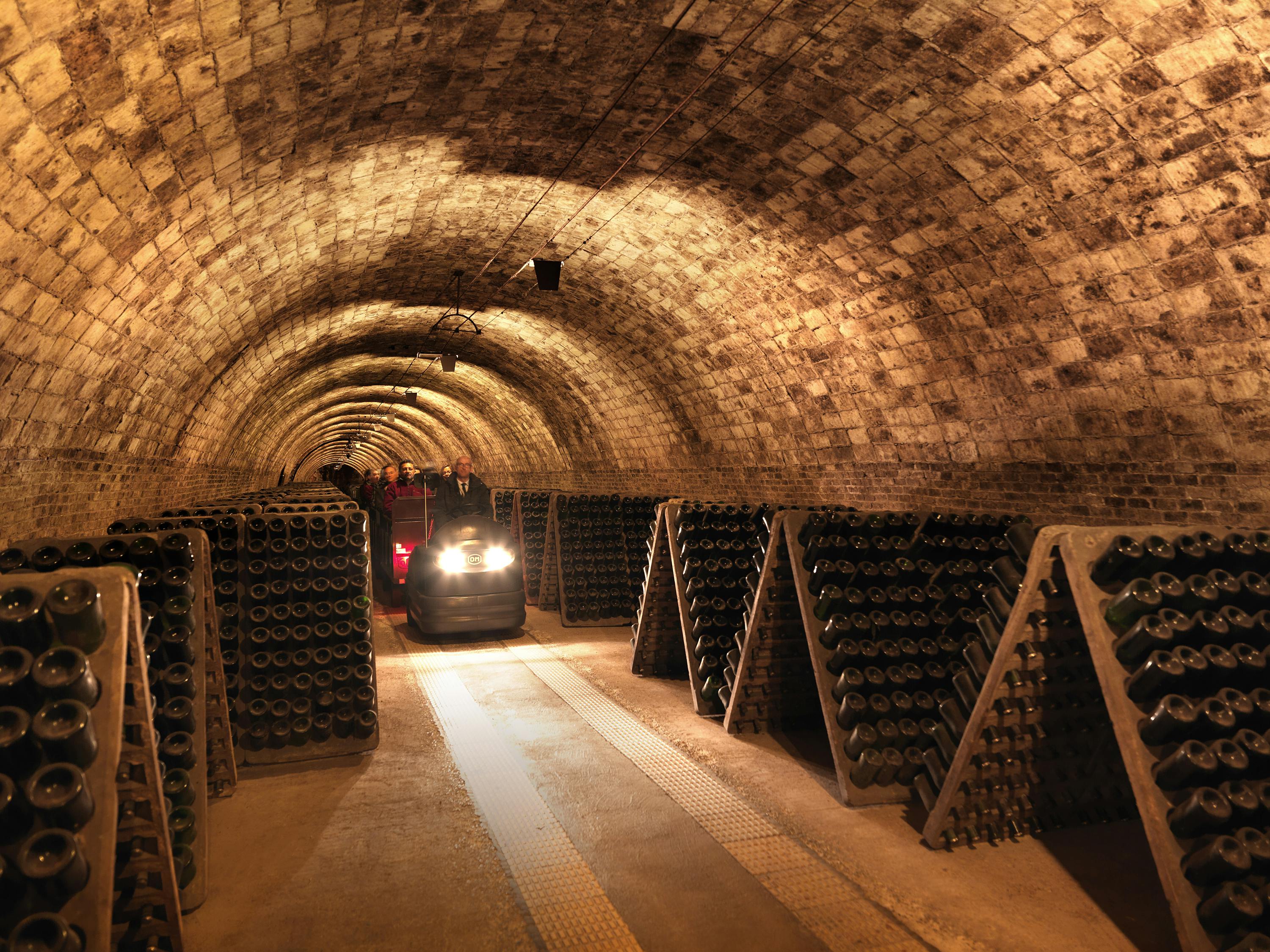 A dimly lit wine cellar with arched ceilings, wine bottles stacked on racks, and a small vehicle with headlights on.