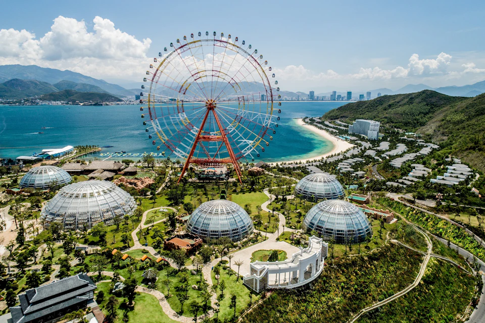A large ferris wheel near a coastal area with domed buildings, green spaces, and a city skyline in the distance.
