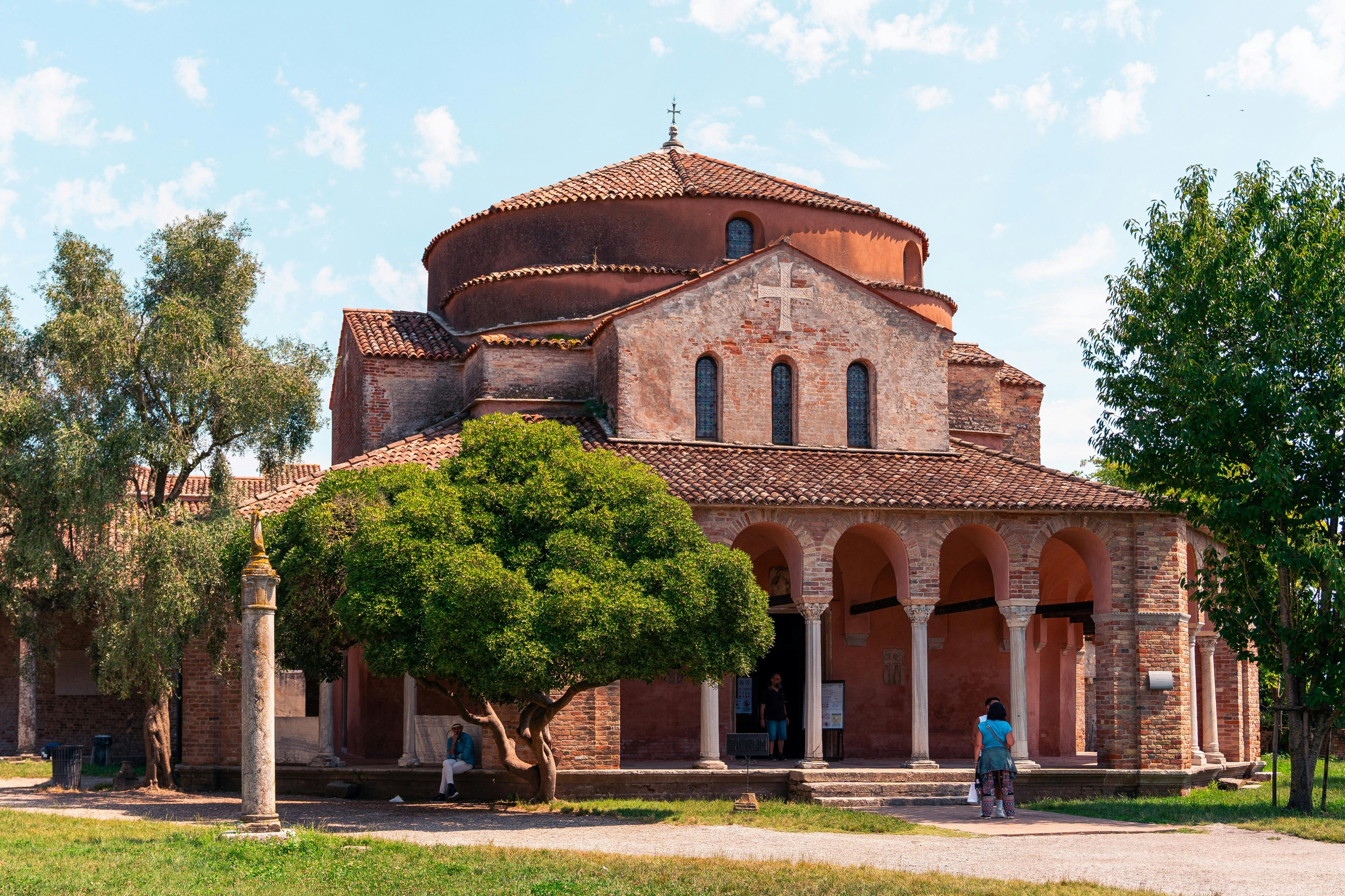 A historic brick church with arched entrances and a large tree in front. Two people standing near the entrance.