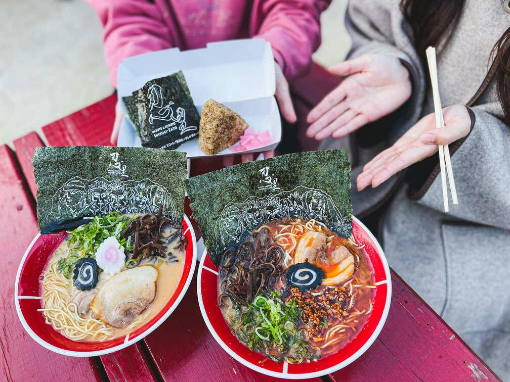 Two bowls of ramen with seaweed, toppings, and broth on a red table, alongside a rice ball in a box held by a person.