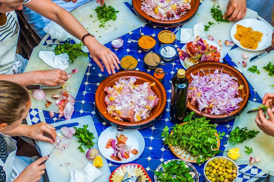 Several people preparing food with chopped herbs, spices, and sliced onions on a blue-tiled table.