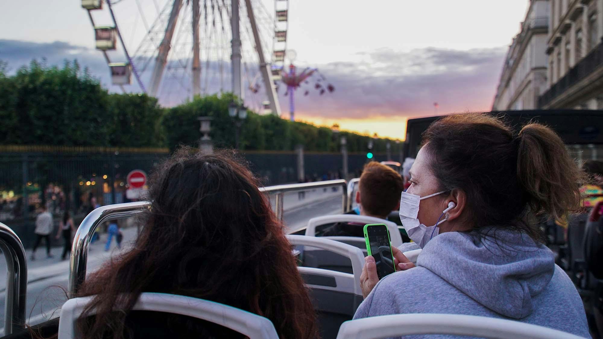 Ludzie w autobusie z otwartym dachem, noszący maskę i patrzący na smartfona, z diabelskim młynem i zachodem słońca w tle.