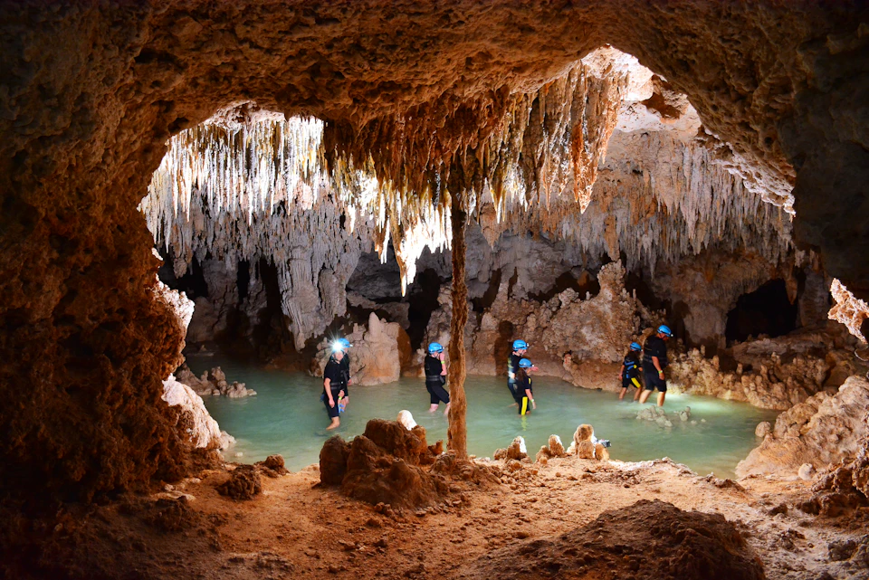 People wearing helmets and headlamps walk through shallow water inside a cave with stalactites and stalagmites.