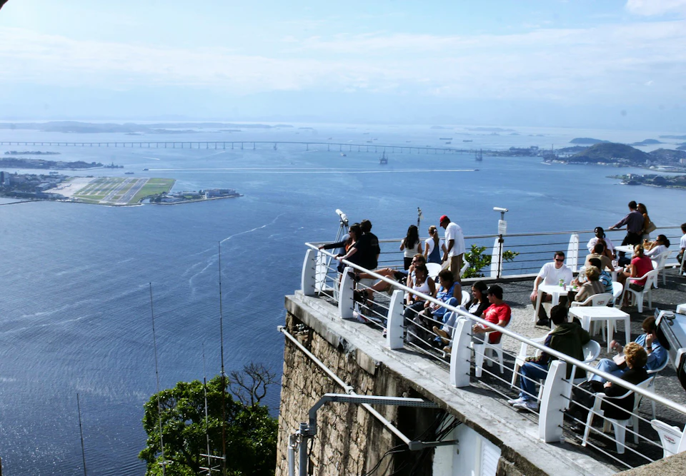 People on a viewing platform overlooking a large body of water with a distant bridge and small greenery-filled island.
