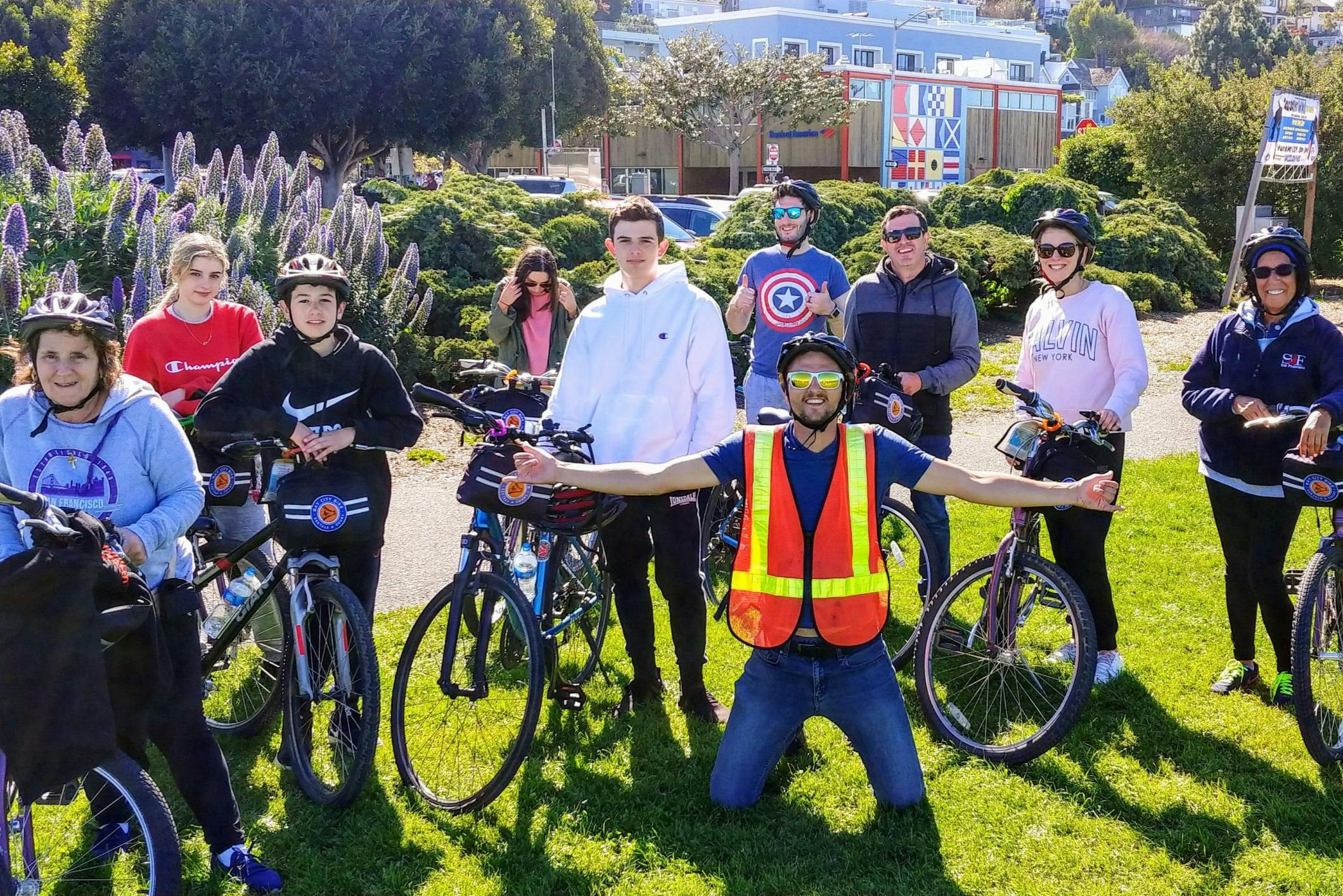 En grupp människor med cyklar, några med hjälm och en person i en neonfärgad säkerhetsväst poserar utomhus bredvid lila blommor.