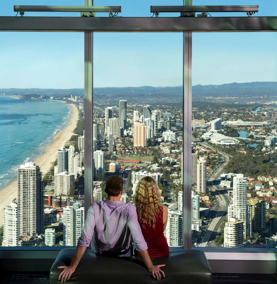 A man and woman sit together, looking out a large window at a cityscape with tall buildings and a coastline.