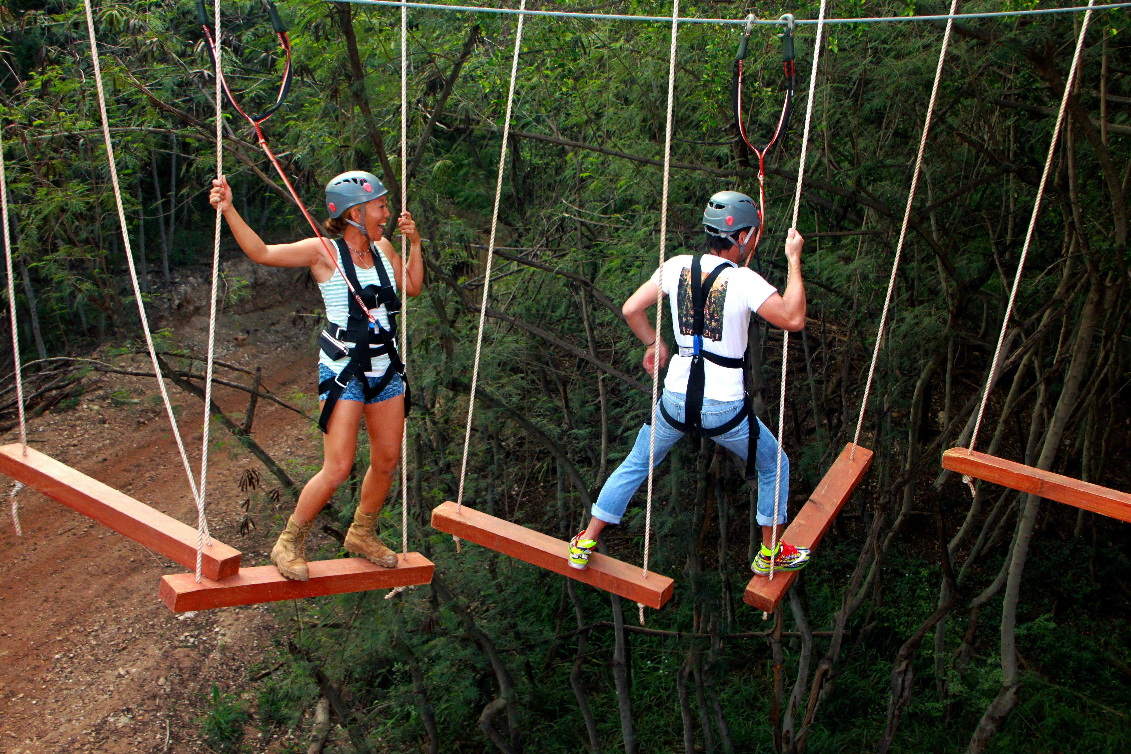 숲이 우거진 지역에서 헬멧과 마구를 쓴 두 사람이 나무 널빤지가 있는 밧줄 다리를 건너고 있습니다.