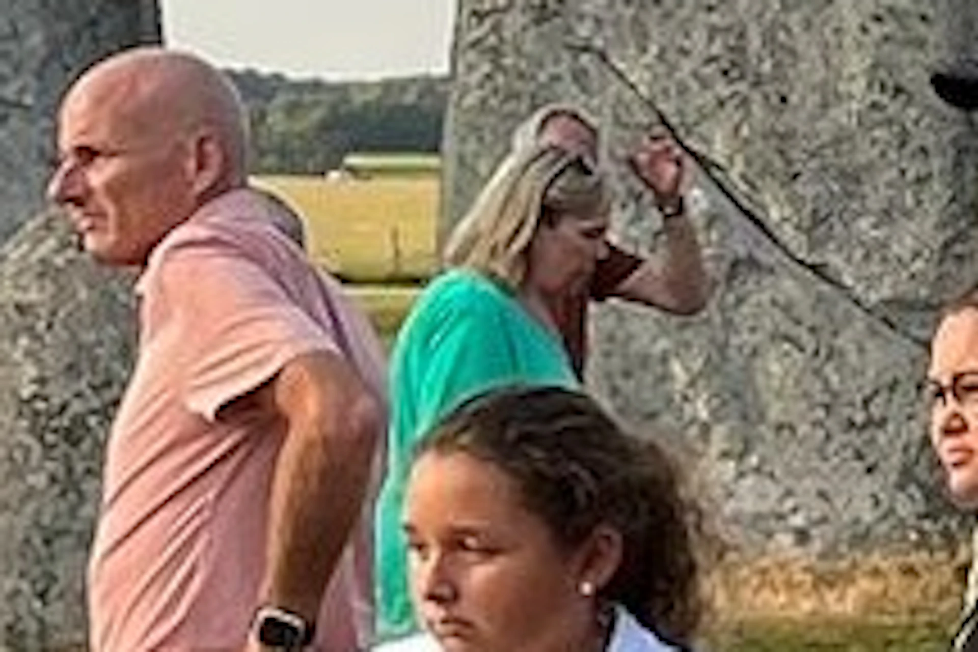 A group of people, including a guide, stand near the Stonehenge monument under a partly cloudy sky.