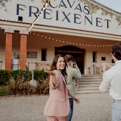 Människor som står utanför en byggnad märkt "Cavas Freixenet", en kvinna som ler och gestikulerar medan andra tar bilder.