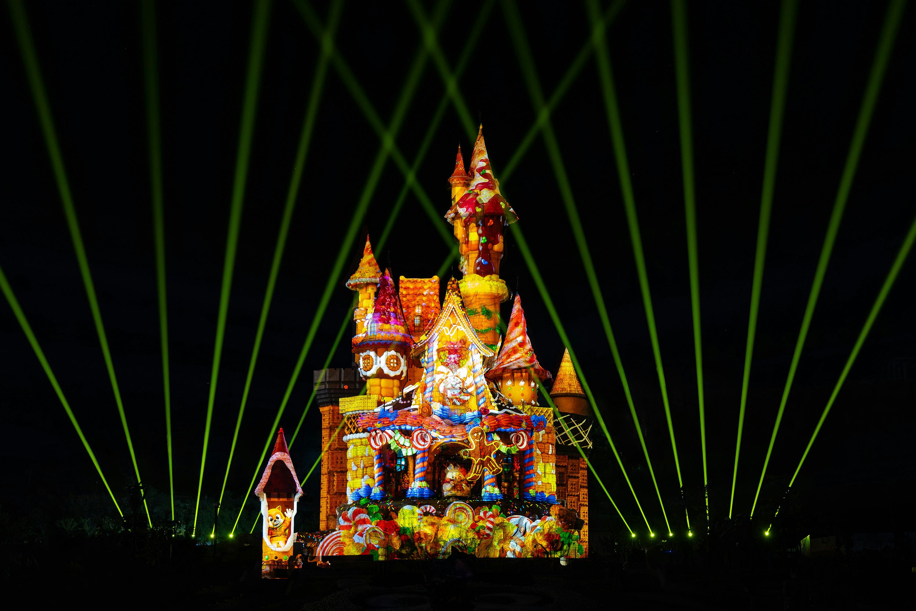 A colorful castle illuminated with holiday-themed projections and green light beams in a dark nighttime setting.