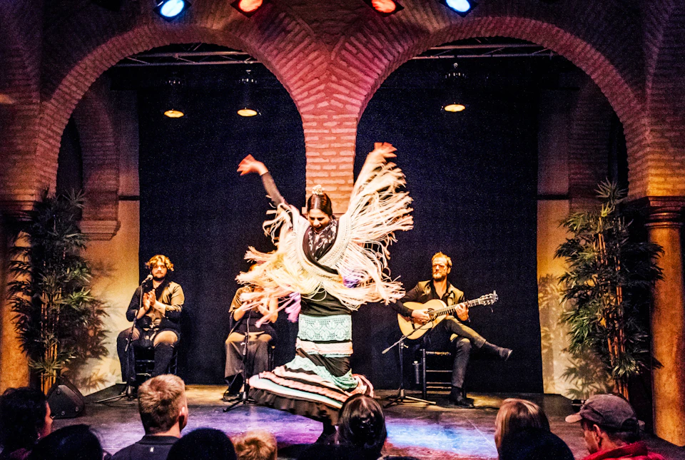 A flamenco dancer performs on stage with musicians seated behind her, one playing a guitar. Arched brickwork forms the backdrop.