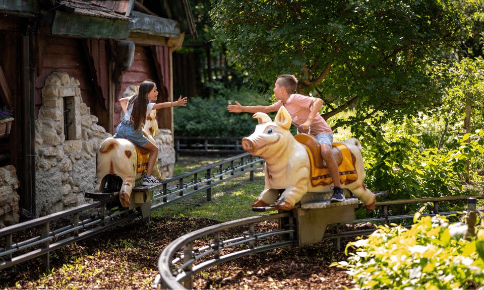 Two people on pig-themed amusement rides reach out to each other, surrounded by greenery and a rustic building.