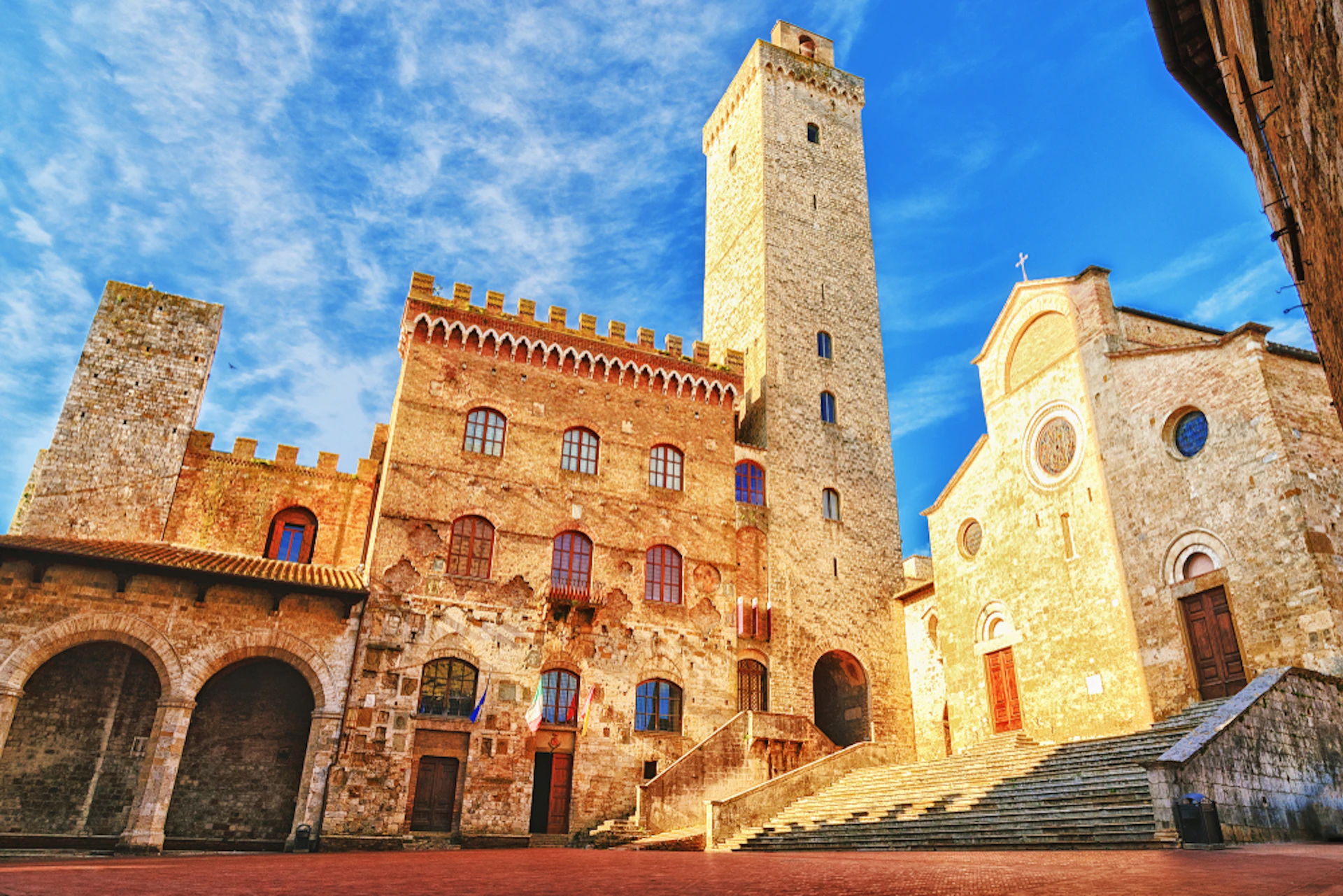 Historic stone buildings with arched windows and a tall tower under a blue sky; broad steps leading to a church entrance.