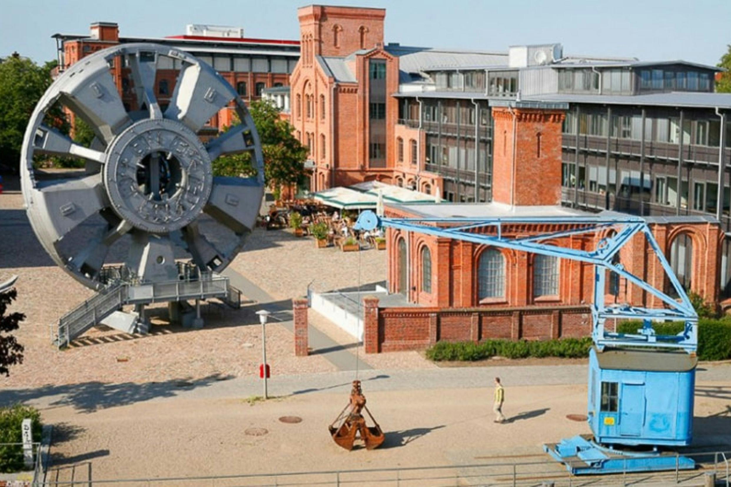 Nádvoří s budovami průmyslového dědictví, velkou kovovou konstrukcí kol, modrým jeřábem a chodící osobou.