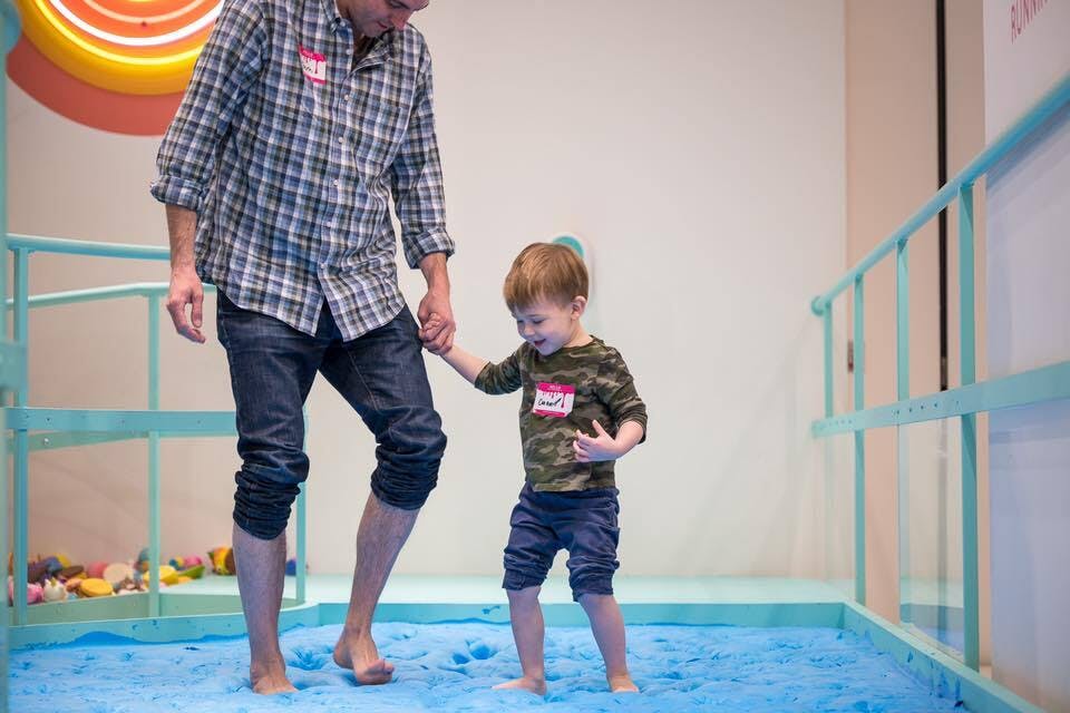 Adult in a checkered shirt and child in camo shirt hold hands, walking barefoot on a blue padded floor. Both wear name tags.