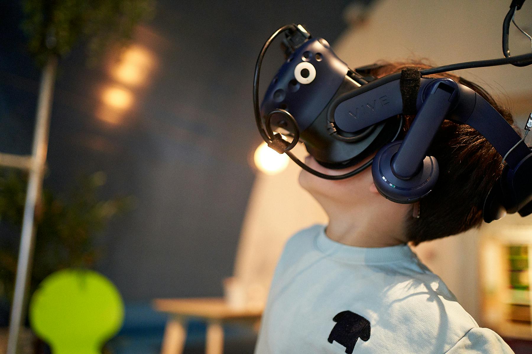 Enfant portant un casque de réalité virtuelle, regardant vers le haut. Fond flou avec éclairage intérieur et éléments de mobilier vagues.