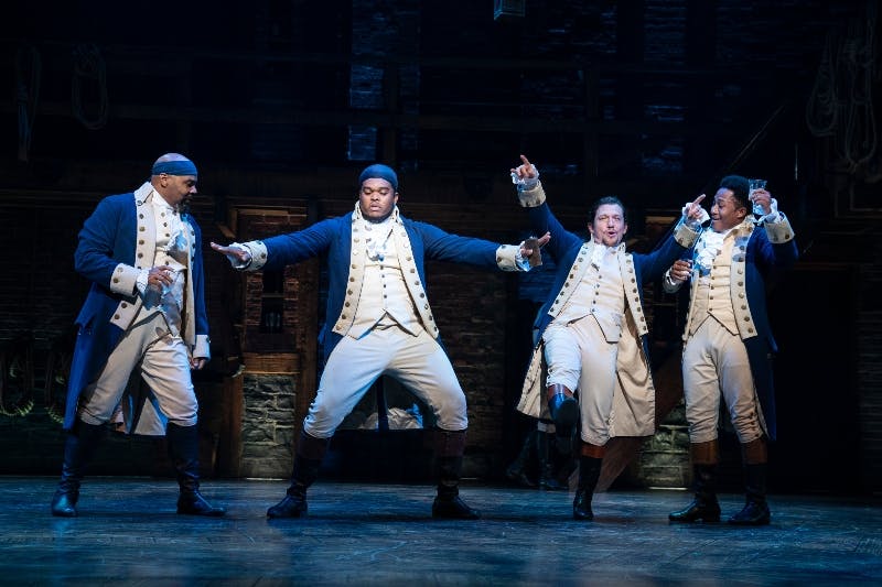 Three men in colonial costumes perform energetically on a dimly lit stage with a brick backdrop.