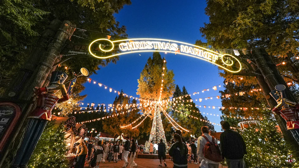 A festive Christmas market at night with a lit-up sign, a decorated tree, and people walking around.