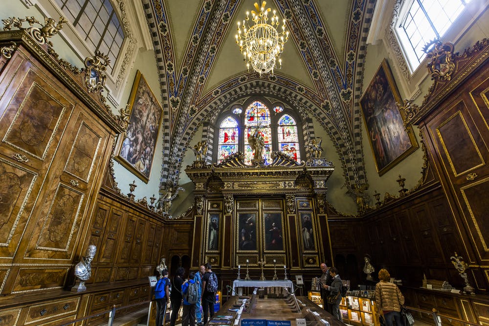 A group of people stands in an ornate chapel with high vaulted ceilings, stained glass windows, and large paintings on the walls.