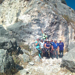 Grupp med åtta personer som bär klätterutrustning och hjälmar vid foten av en stenig klippa under en klarblå himmel.
