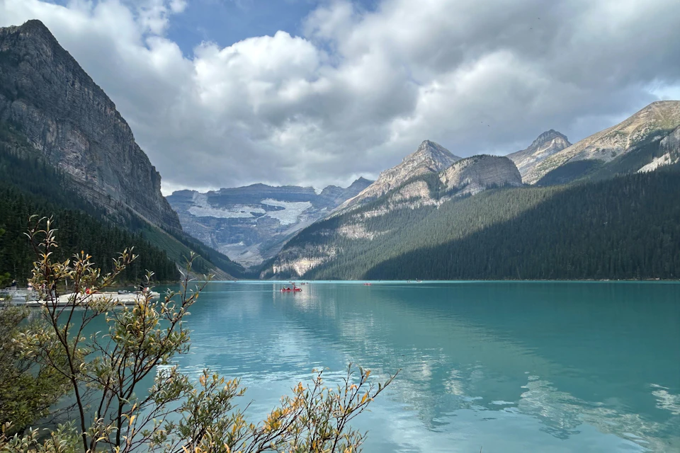 A turquoise lake surrounded by mountains under a cloudy sky; a few people in red canoes are visible on the water.