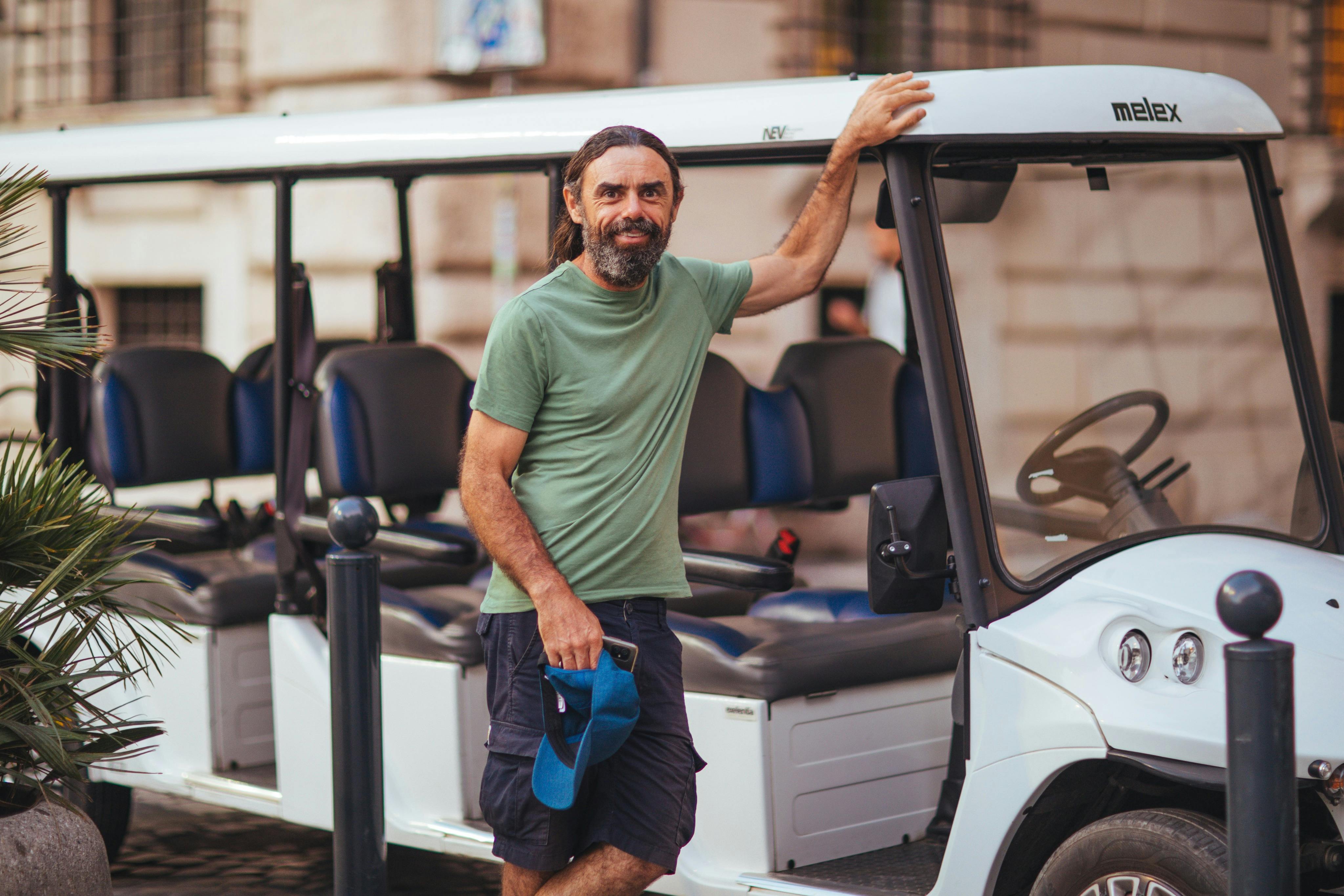 A man in a green shirt and shorts stands beside a white tram-like vehicle, holding a blue cap and smiling.
