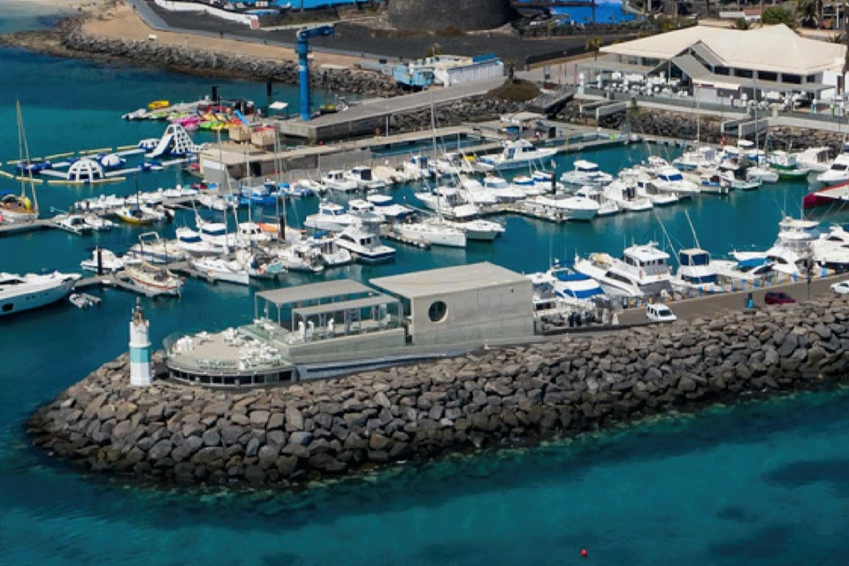 Aerial view of a marina filled with boats, adjacent to rocky breakwaters and a modern building near turquoise waters.