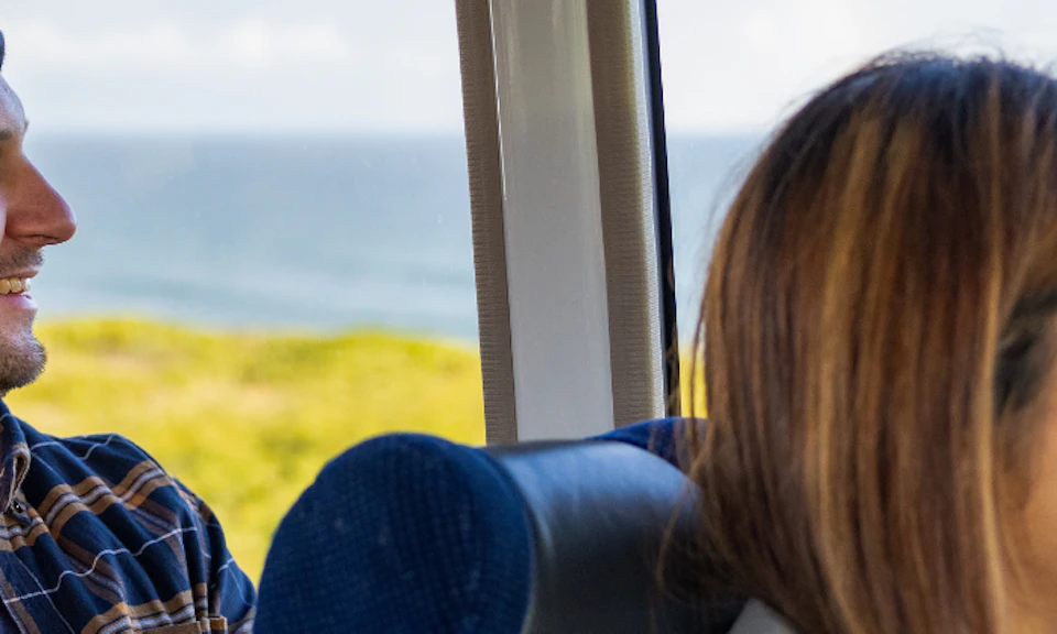 A man and a woman are sitting on a bus, smiling and looking out the window at a scenic landscape with greenery and ocean.