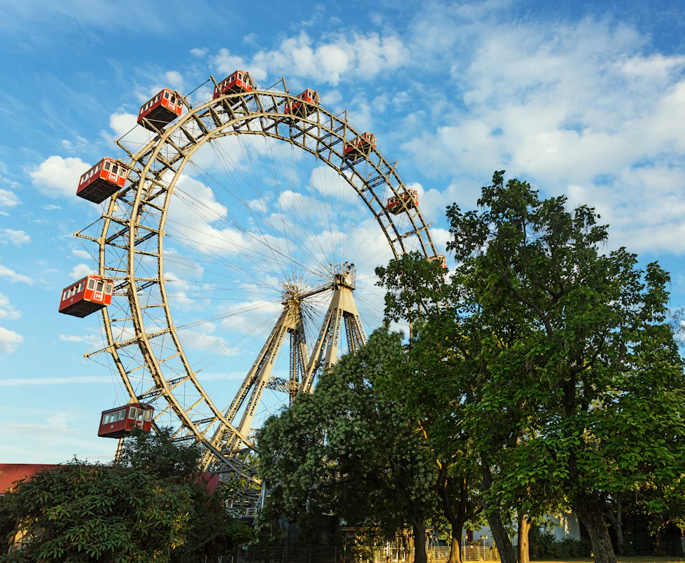 Large Ferris wheel with red cabins under a blue, partly cloudy sky, surrounded by lush green trees.