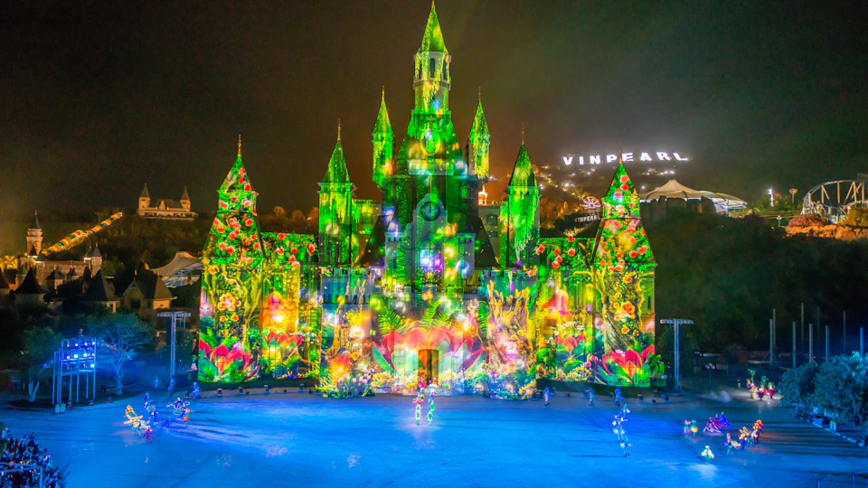 A brightly lit castle with colorful projections at night, featuring floral and nature themes. "Vinpearl" sign in background.