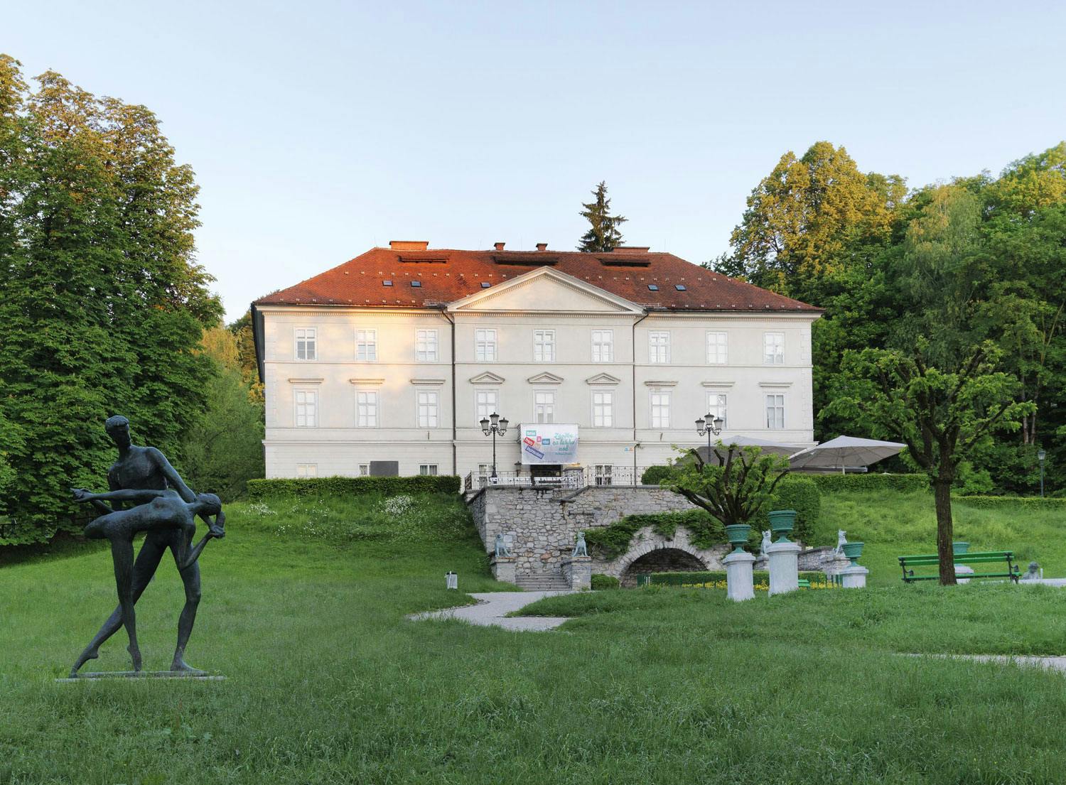 Historické bílé sídlo s červenou střechou, obklopené zelení, kamenným mostem a sochou v parku.