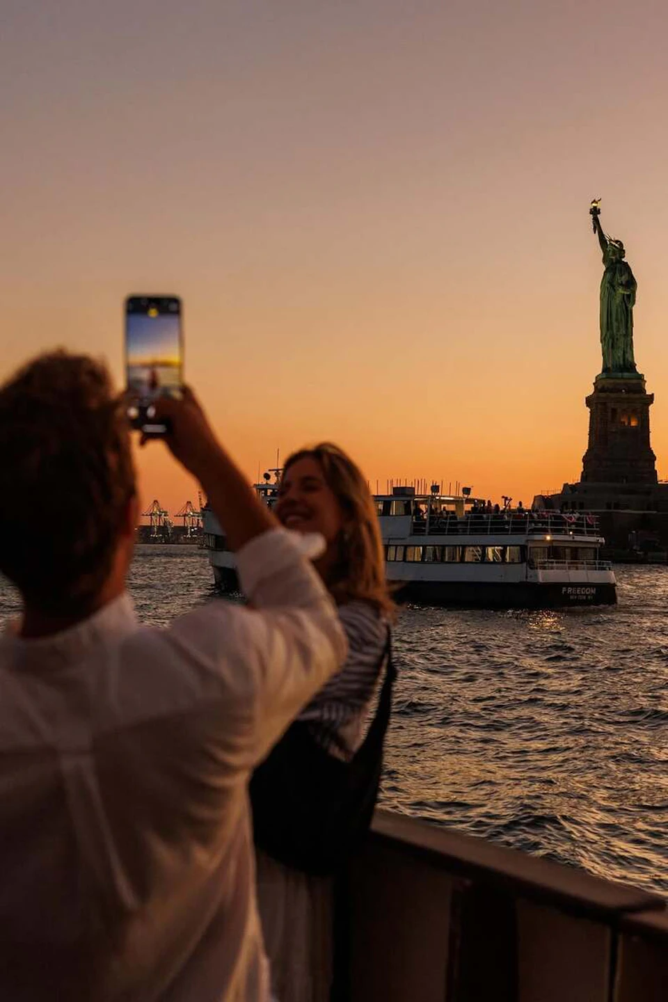 Two people at sunset near a harbor, one taking a photo of the Statue of Liberty with a smartphone, with a ferry in the background.
