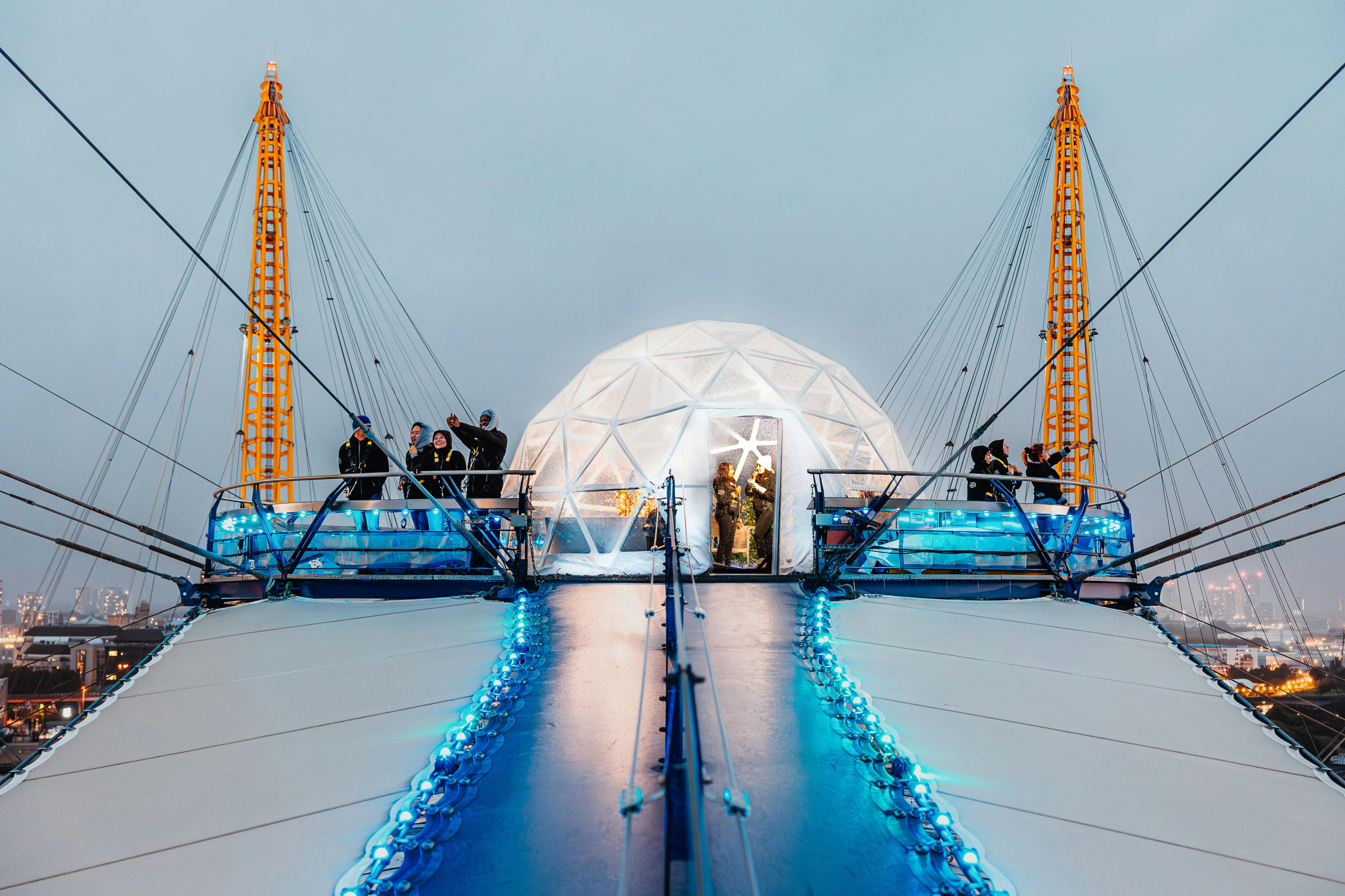 People gather on a modern rooftop platform with blue lights and a white geodesic dome, surrounded by yellow support structures.