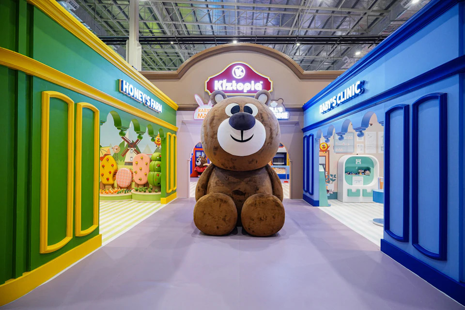 A large teddy bear sculpture sits in the center of a colorful indoor play area with themed storefronts on either side.