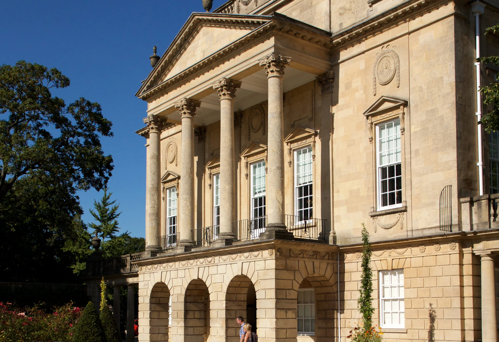 A grand building with tall columns, arched windows, and a decorative facade. Two people are walking near the entrance.