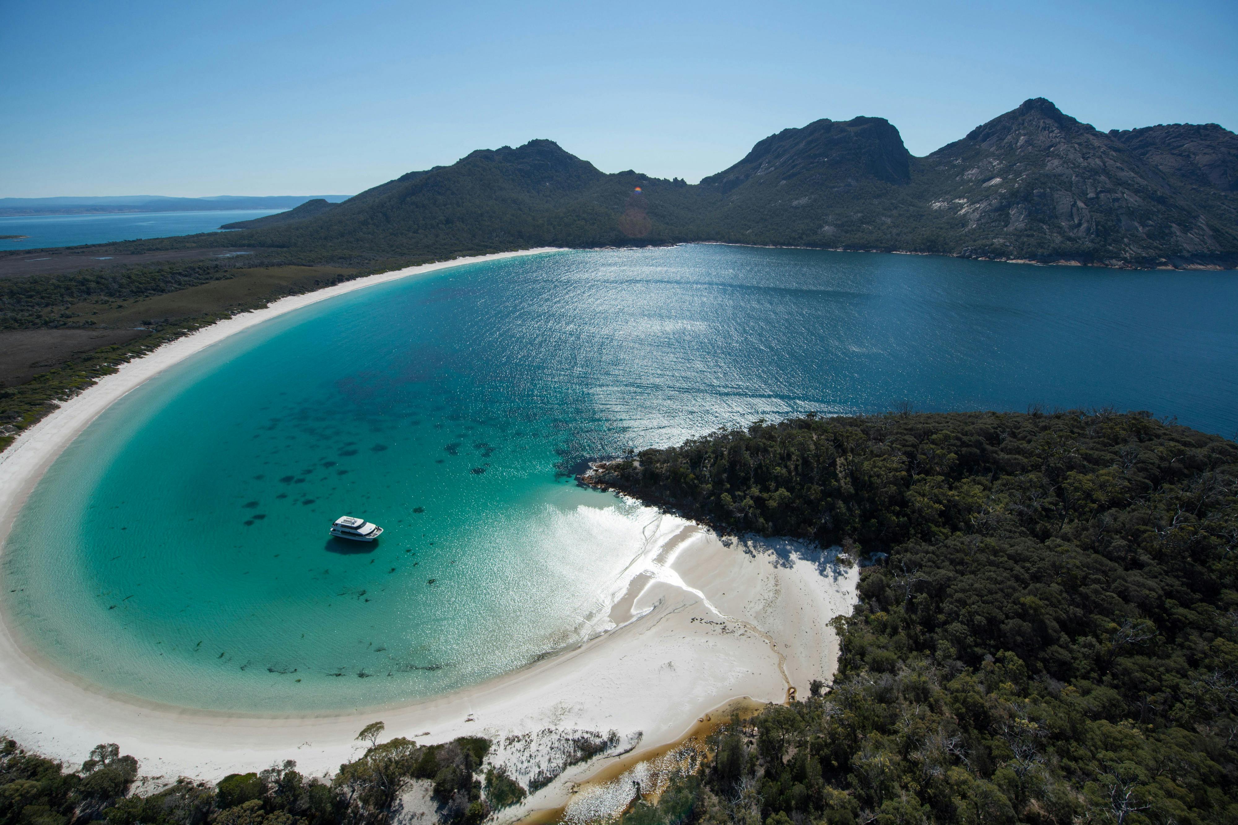 Aerial view of a bay with turquoise water, a sandy beach, a forested area, and a boat anchored near the shore.