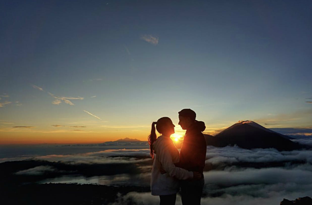 Pár stojí těsně u sebe na vrcholu hory při východu slunce, s mraky, vzdálenými horami a jasnou oblohou v pozadí.