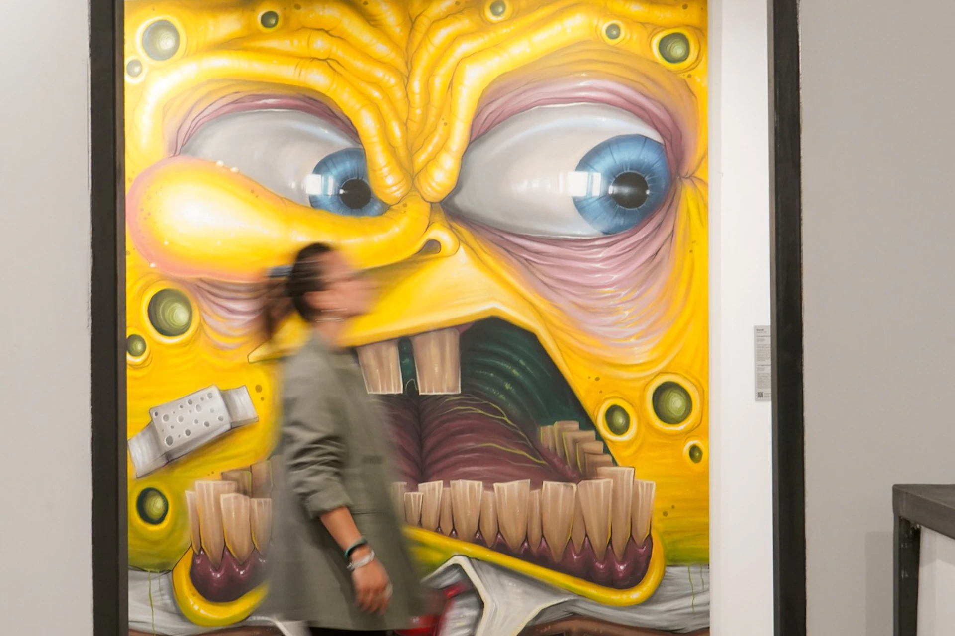 A person walking past a colorful, exaggerated cartoonish mural with a yellow face and large, dramatic facial features.
