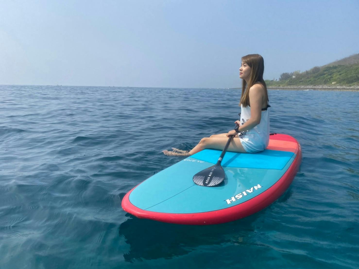 A woman sitting on a red and blue paddleboard holding a paddle, floating on calm blue water near a shoreline.
