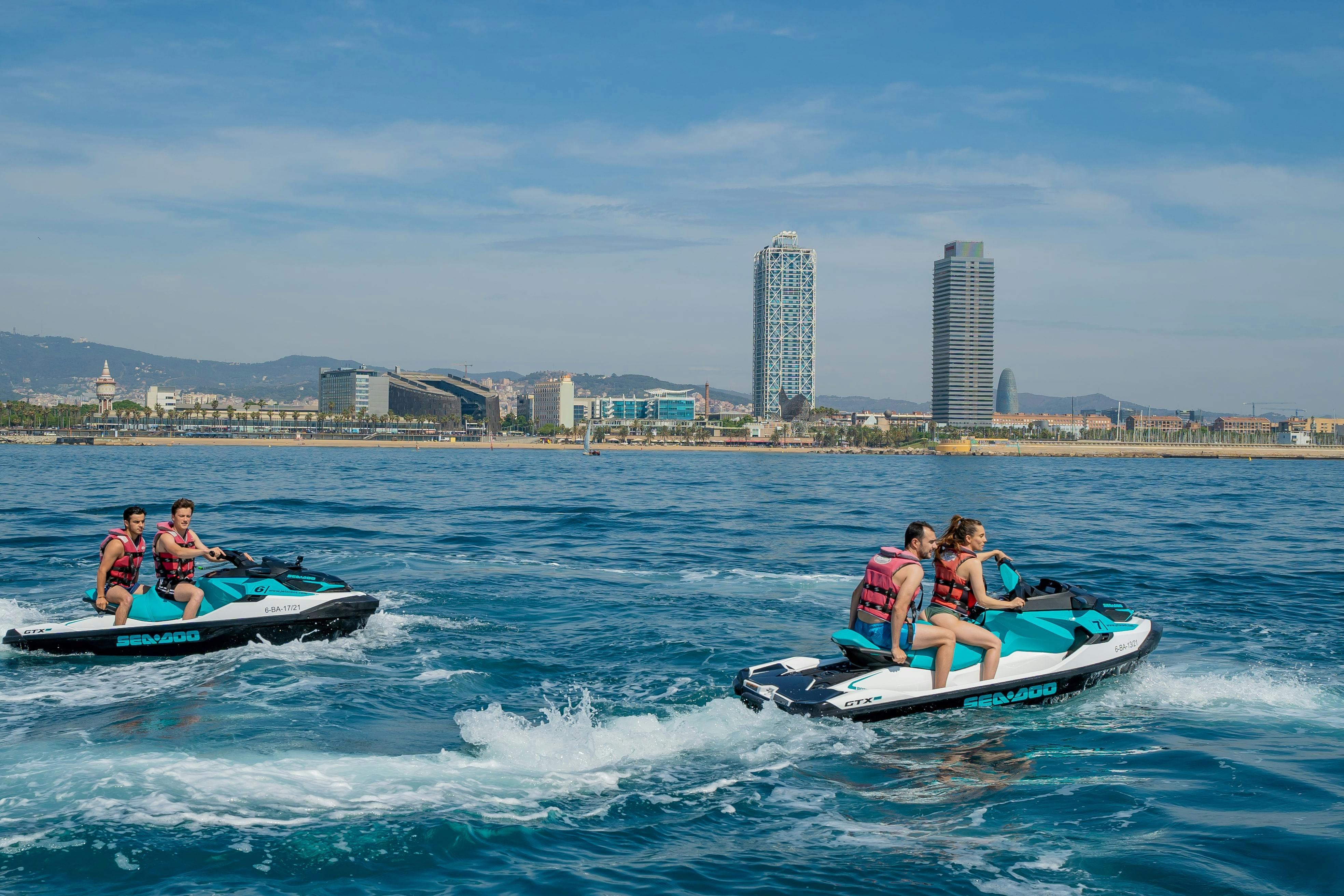 Two pairs of people ride jet skis on the water near a city shoreline with tall buildings, under a clear blue sky.