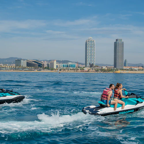 Två par människor åker vattenskoter på vattnet nära en stadsstrand med höga byggnader, under en klarblå himmel.