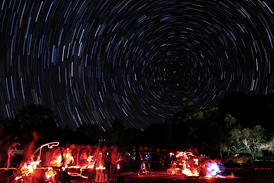 A long exposure night photo showing star trails in the sky and illuminated objects on the ground near trees.