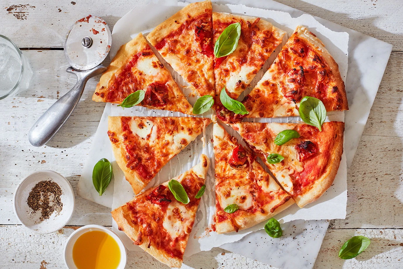 A sliced margherita pizza with basil leaves, next to a pizza cutter and small bowls of yellow sauce and olive oil on a white surface.