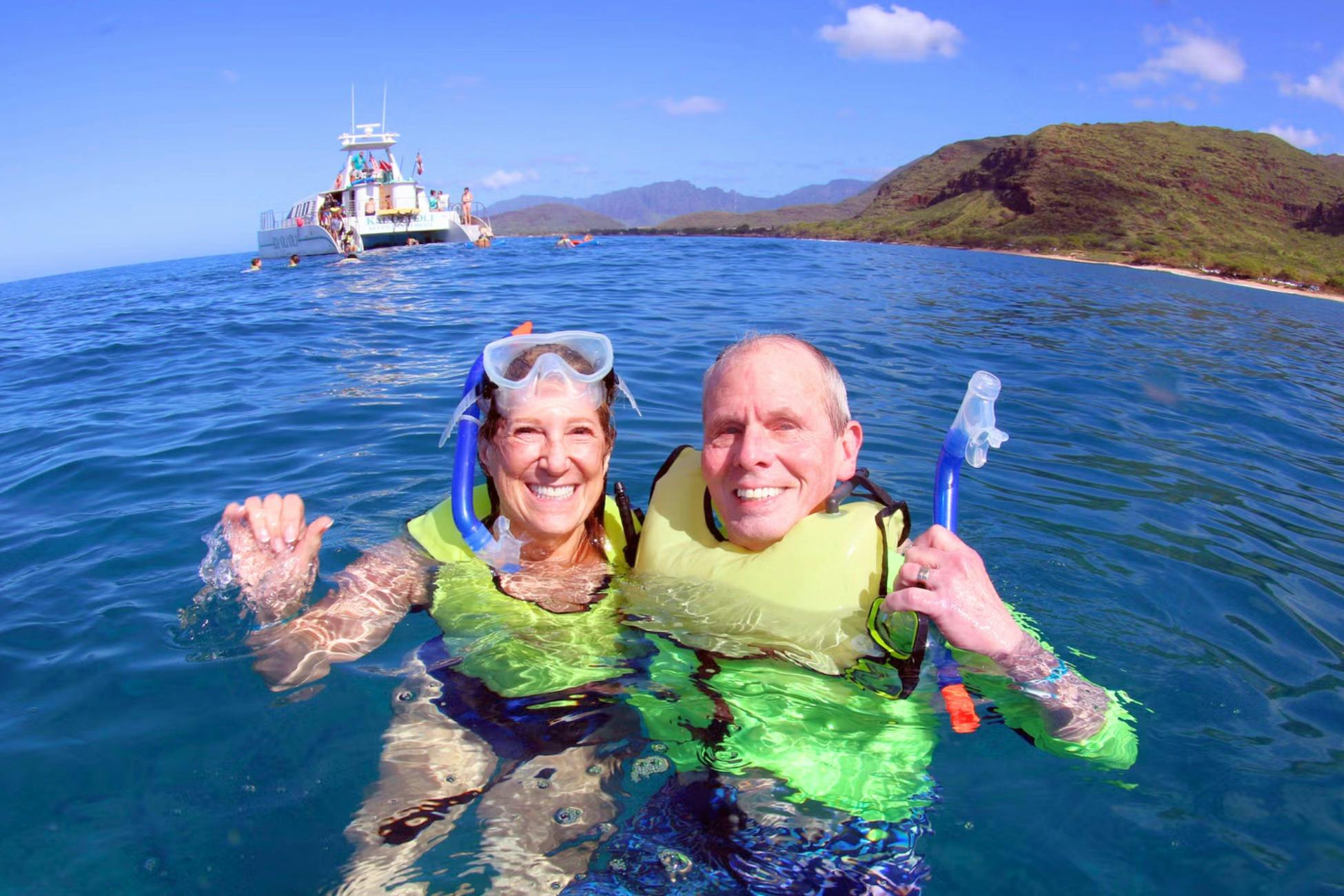 Two people wearing snorkeling gear and life vests in the ocean, with a boat and mountainous coastline in the background.