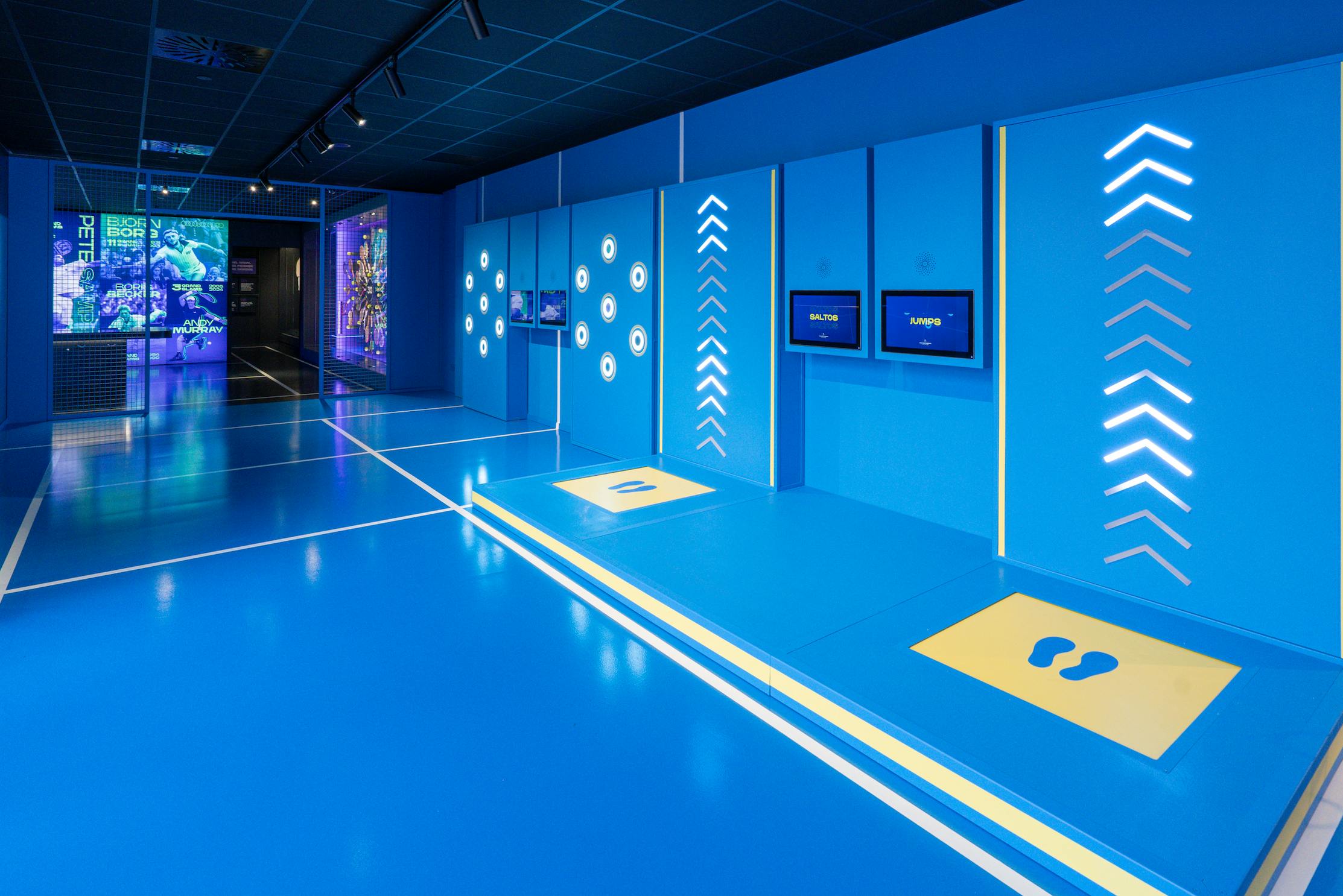Una moderna exposición interactiva con paredes azules, pantallas y marcas en el suelo, con flechas y huellas iluminadas.