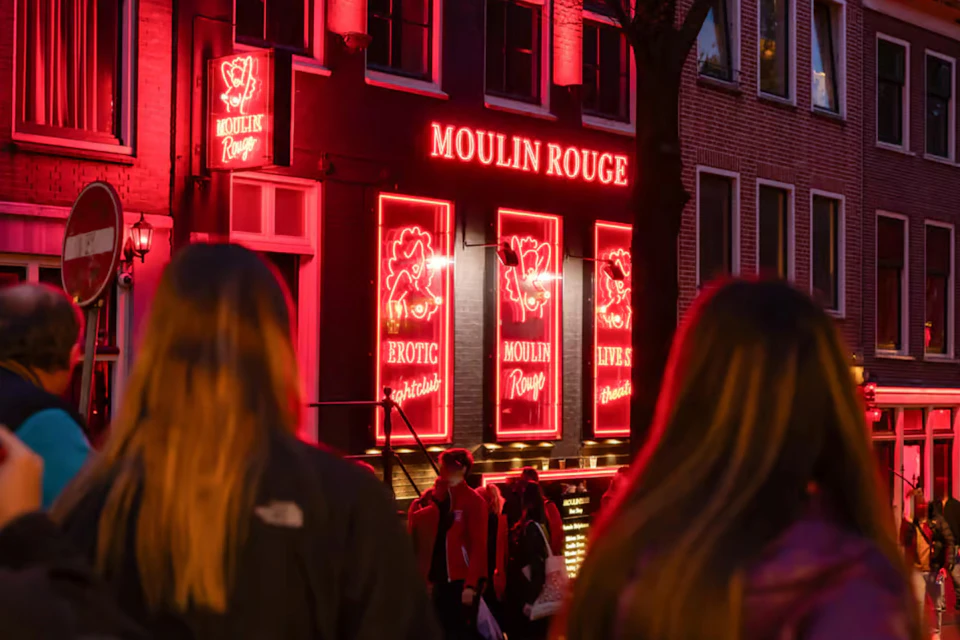 People walking past a building with bright red neon signs reading "Moulin Rouge," advertising erotic and live shows.