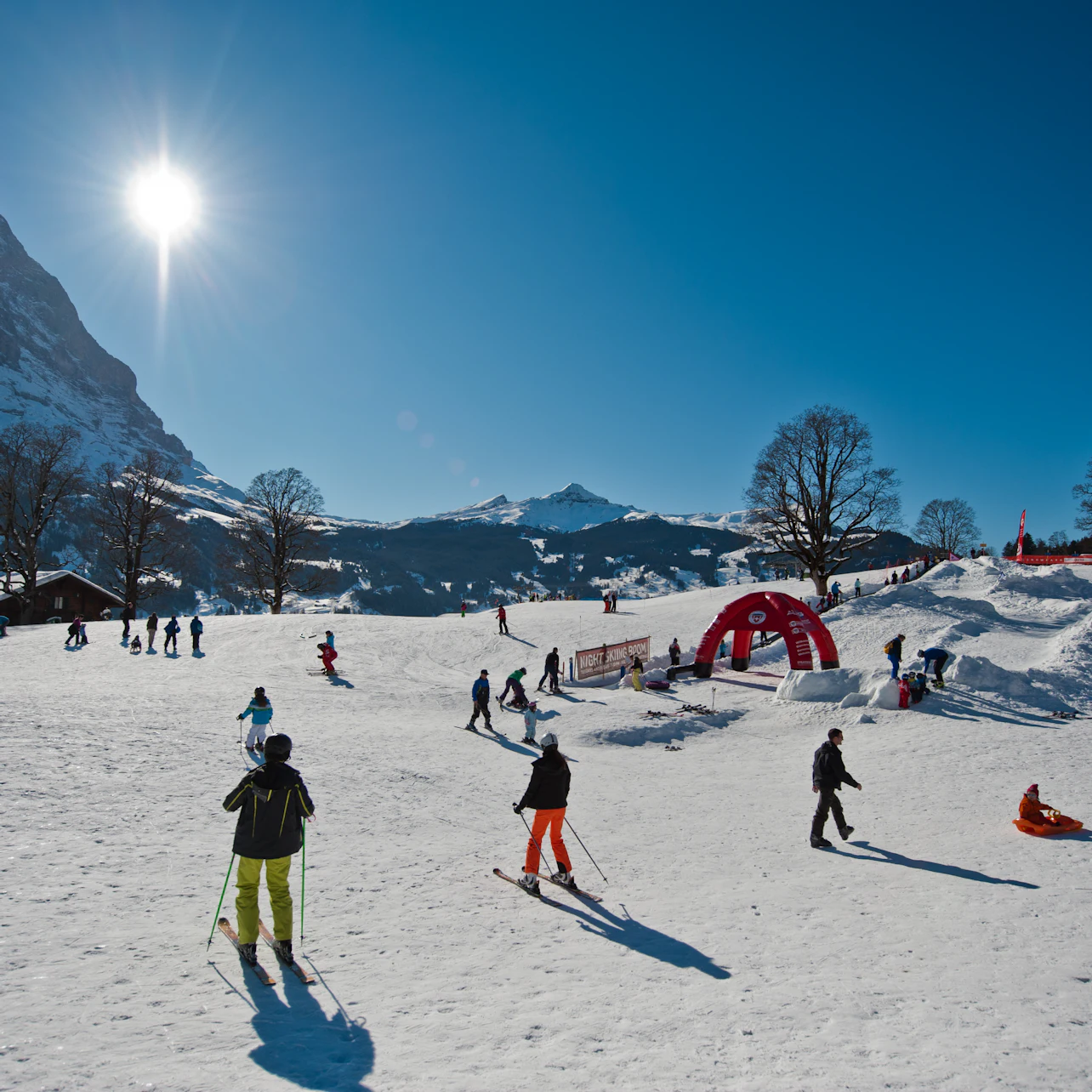 Skiers and snowboarders enjoy a sunny day on a snowy slope with mountains and clear blue sky in the background.