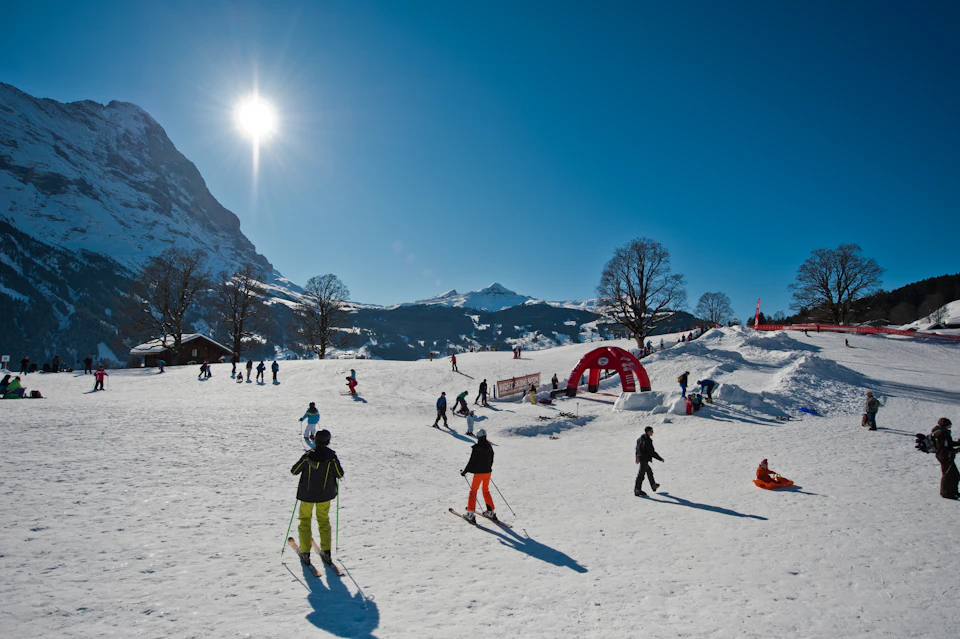 Skiers and snowboarders enjoy a sunny day on a snowy slope with mountains and clear blue sky in the background.